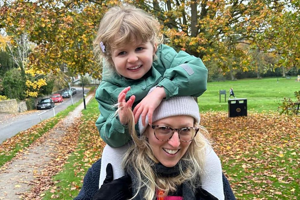 Megan, who is wearing a grey beanie, glasses and has blonde hair, is holding her daughter Joy on her shoulders. Joy is wearing an emerald green coat and is also smiling. They are standing outside in a grassed area. The picture looks very autumnal.