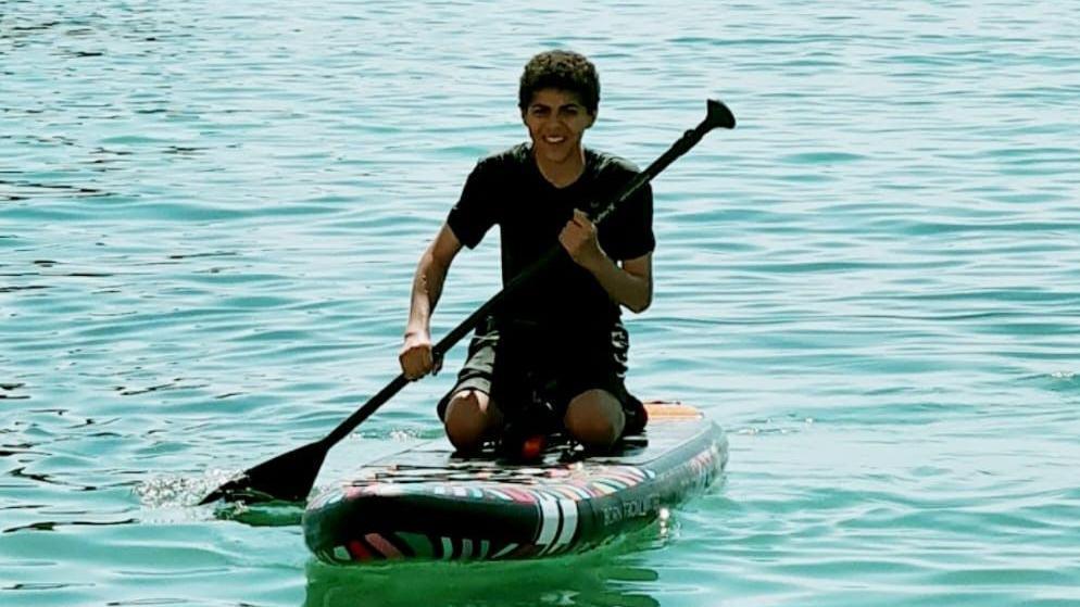 Zak is kneeling on a paddleboard on bright blue water. He is laughing as he paddles through the water.