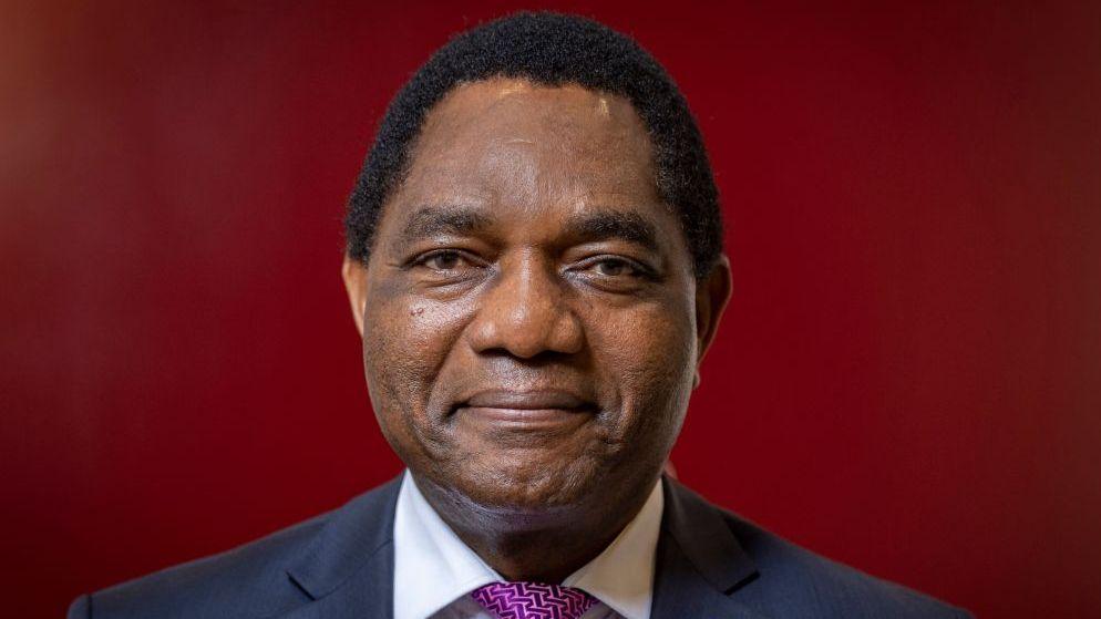 Zambian President Hakainde Hichilema smiles at the camera, wearing a suit and a patterned purple tie.