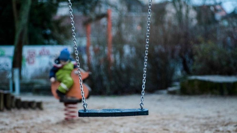 A generic image showing a child's swing in the foreground. A child, who is blurred on the photo and wearing a blue hat, is in the background sitting down.