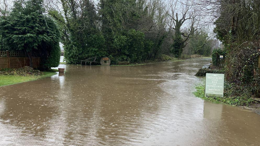 Water floods a road in North Yorkshire, a sign for Brickfields Farm can be seen as well as a milllstone with a sign on it 