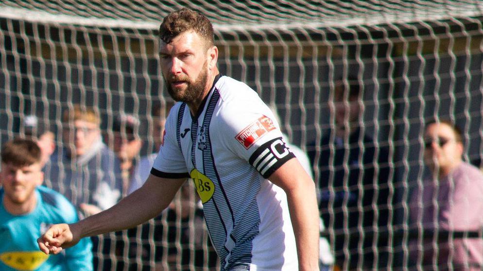 Scott Arnold in a white Wimborne Town shirt wearing the captains armband too playing on the pitch