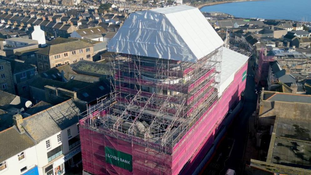 Aerial image showing the building surrounded in scaffolding, with pink netting or white covering on top in some places. The rooftops of surrounding buildings can be seen