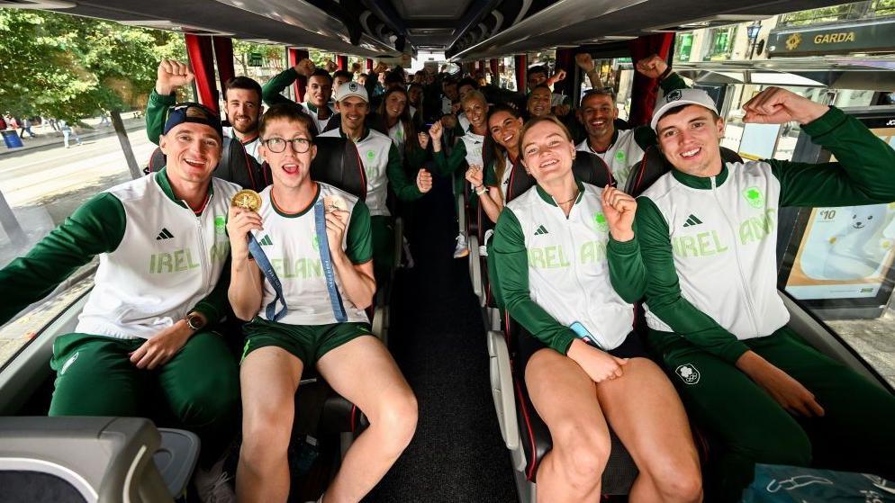 The Ireland Olympic team arrive in a coach wearing Ireland tops and some holding up medals 