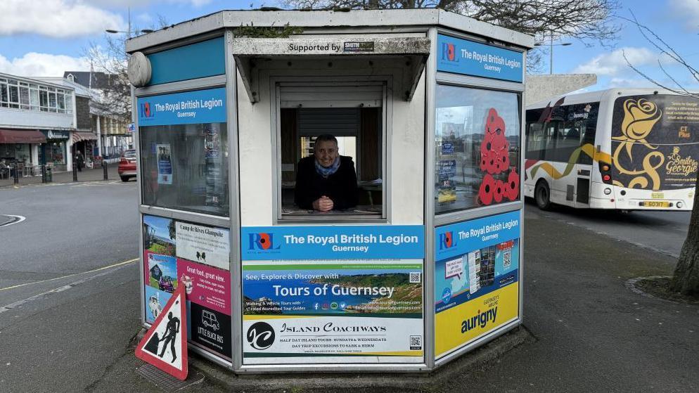 A woman leans over the open hatch of a hexagonal kiosk at a bus terminal. It has the logo of the Royal British Legion on it and a bus is driving by to the right of the kiosk.