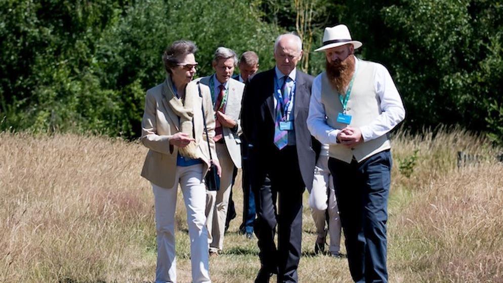 A woman pinned back brown hair, wearing a pale suit and sunglasses with a handbag over her shoulder, walking in a grassy field alongside a man with a white hat and a waistcoat with a large ginger beard. Behind them there are more men in suits