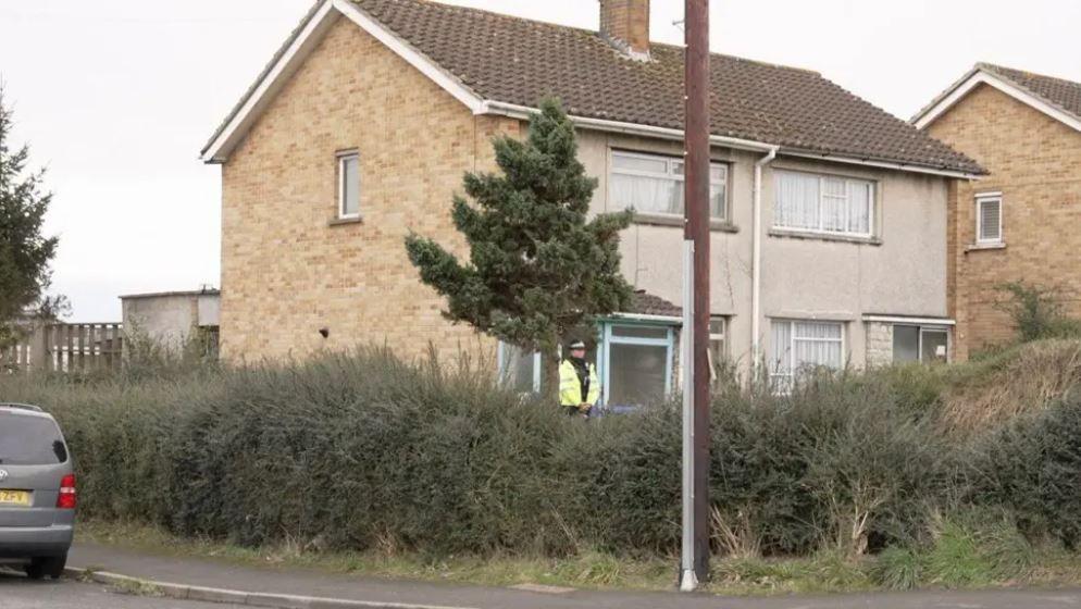 A house on the corner of a residential street. It is a two floor home with sandstone coloured bricks on the side and a small window, and a plastered frontage. There is a police officer guarding the front door. In the foreground there is a large hedge which separates the home from the pavement and road.