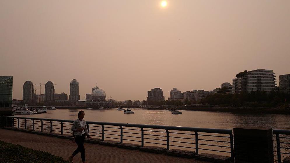 People walk next to the harbour in Vancouver as smoke from wildfires hangs over the city on September 05, 2025. The sky is greyish orange. 