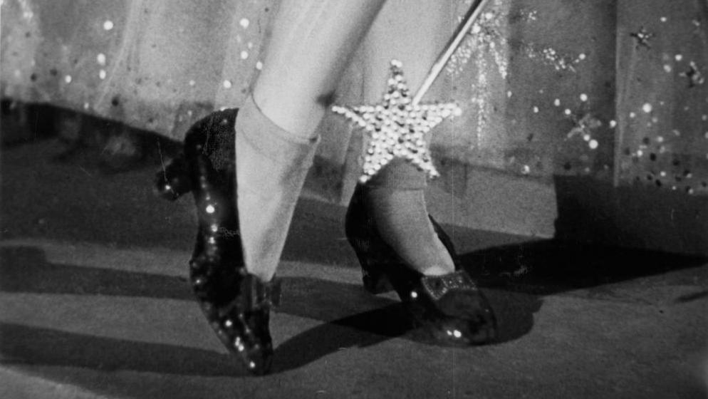 A black and white close-up shot from the 1939 film showing Dorothy lifting up one heel while wearing the shoes, with a star-shaped wand pointing down at them. Glinda's full sequinned net skirt can be seen behind her