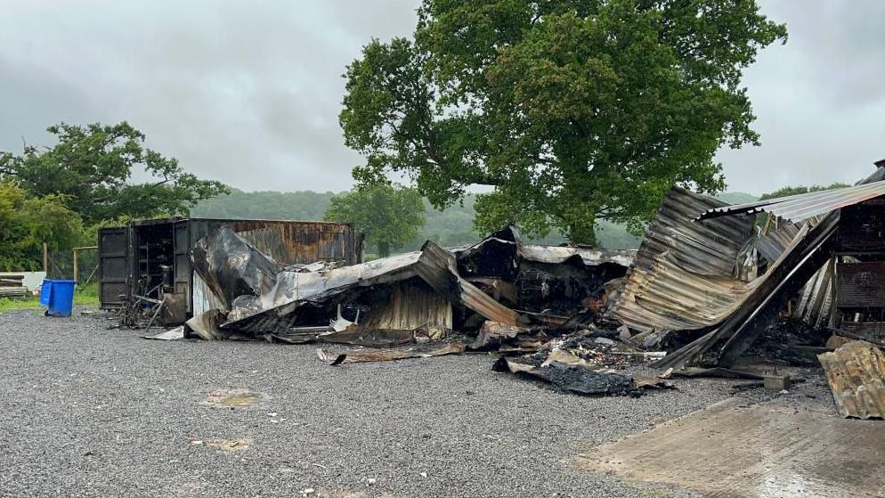 A farm shop building that has been destroyed in a fire. The rubble is now piled up on the floor. A large tree is behind the pile of debris.