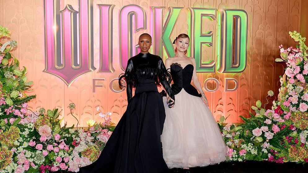 ariana grande and cynthia erivo holding hands on a red carpet