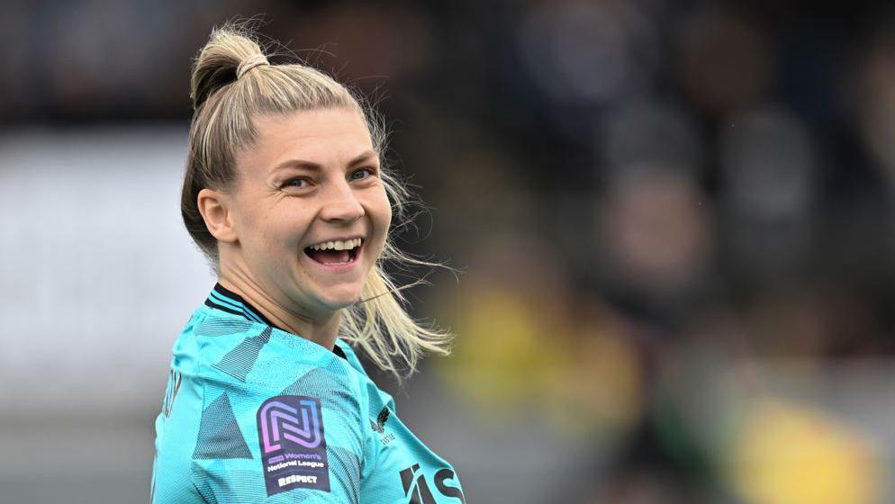 Grace Donnelly. She is wearing a light blue Newcastle United shirt and is standing on a pitch. She has turned her head side-on to the camera and is laughing.