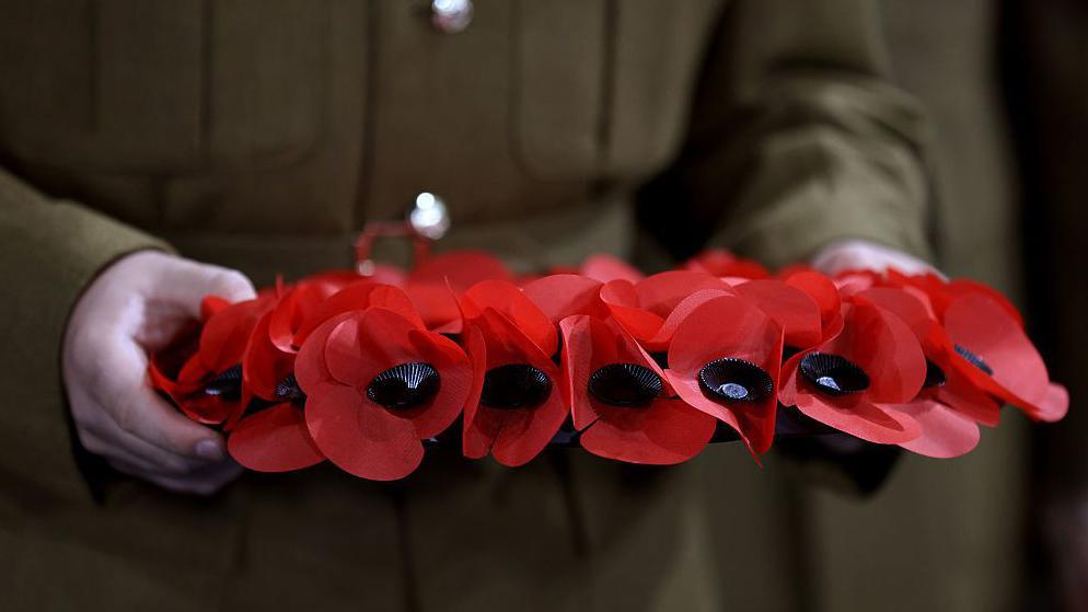 A close up image of a wreath of poppies, being carried in two hands by someone wearing historical military uniform.