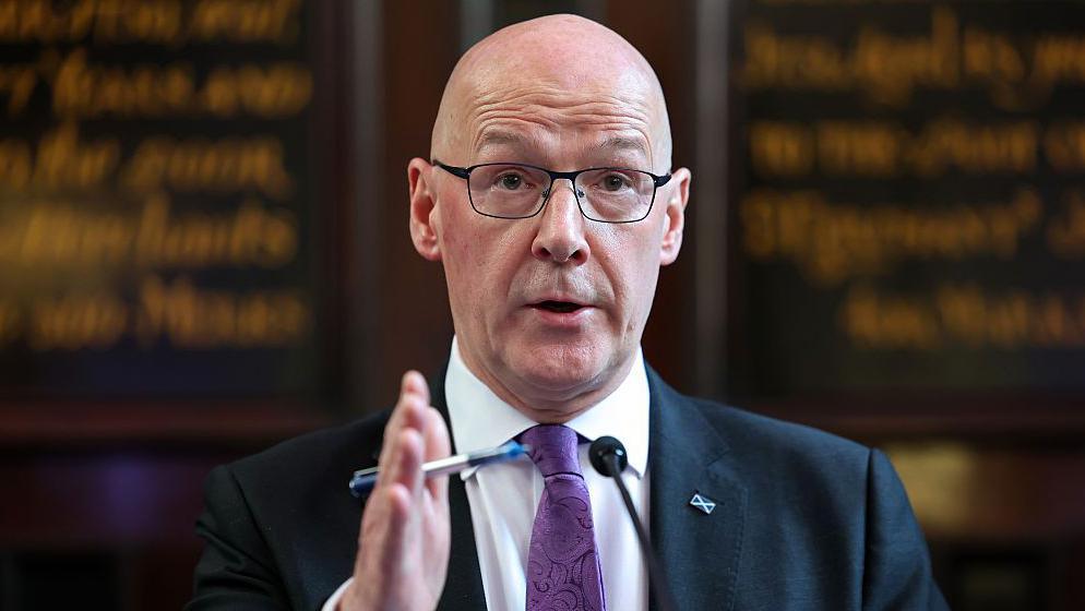 John Swinney, who is wearing a navy blazer, a purple tie and a white shirt, addressing a press conference. Behind him, blurred, is a board with calligraphy writing on it.