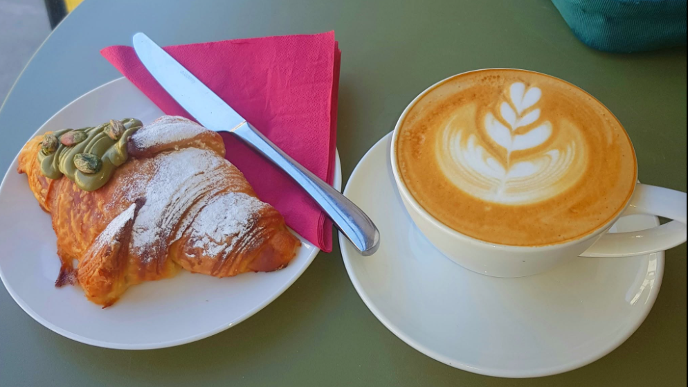 A coffee and pistachio croissant at a cafe. They're both on white plates on a green table. The coffee has latte art on top.