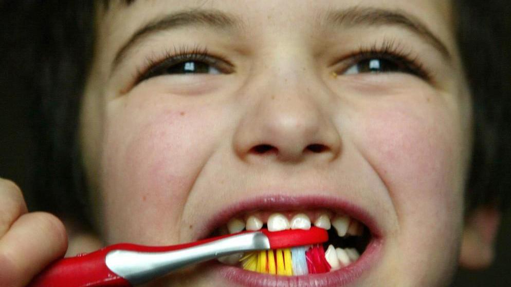 A schoolboy with dark hair and eyes brushing his teeth with a red toothbrush with coloured brushes