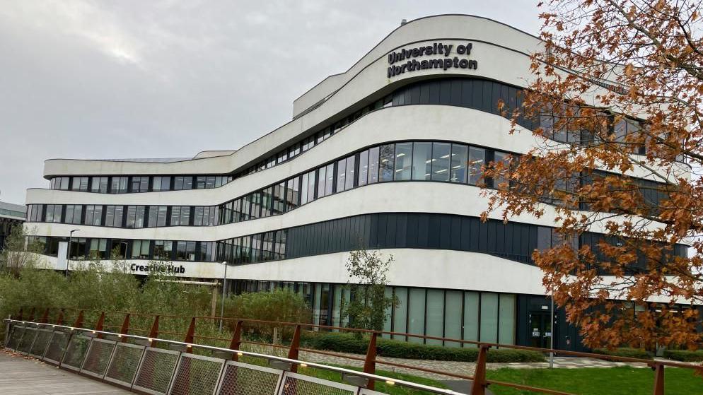 A black-and-white building with lots of glass windows surrounded by grass, grey sky and a tree with brown leaves.