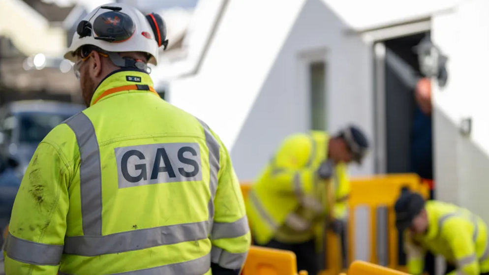 A man wearing a white hard hat and a yellow high viz jacket with GAS written in large capital letters on his back stands facing away from the camera while two other people in high viz clothing work behind a yellow barrier.