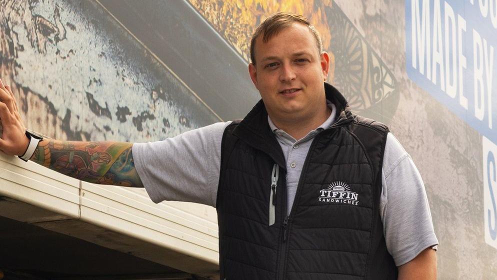 A man with short brown hair wearing a grey polo shirt and black vest. He is leaning against a truck.