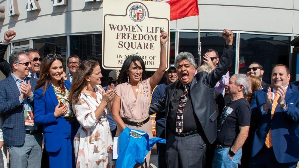 Elham Yaghoubian and Roozbeh Farahanipour - with their fists raised - stand in front of a sign with a group of about 12 other people clapping in support. The sign says - Women Life Freedom Square - in the Westwood neighborhood of Los Angeles.