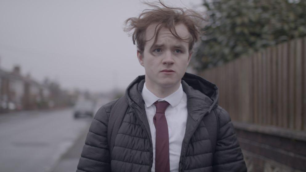 A still from Judah Cousin's film More Than What You See. He is walking down the street wearing his school uniform. He has a sad look on his face. He has a black puffa coat on, white shirt and red tie. His ginger hair flies in the wind.