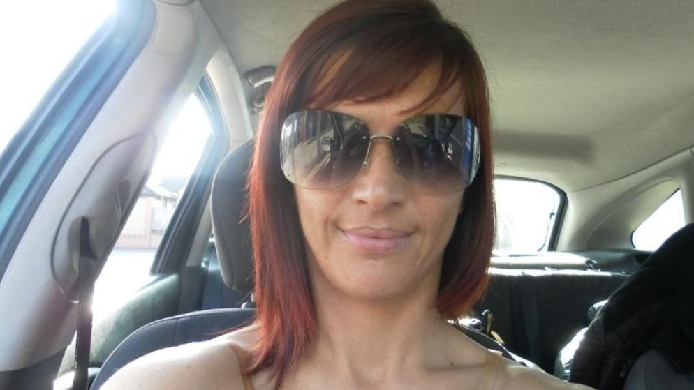 A woman with red shoulder length hair and large sunglasses looks at the camera for a selfie in the passenger seat of a car