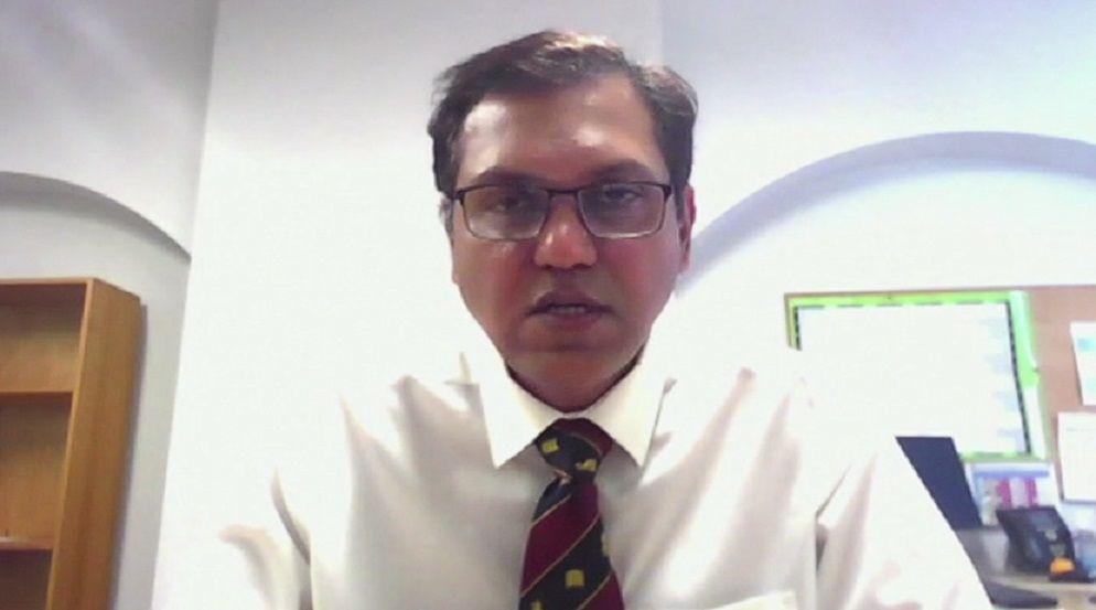Hemant Nemade with medium-length dark hair and glasses. He is wearing a white shirt and red and blue tie and looking at the camera. There is a bookshelf to the left behind him and a noticeboard to the right.