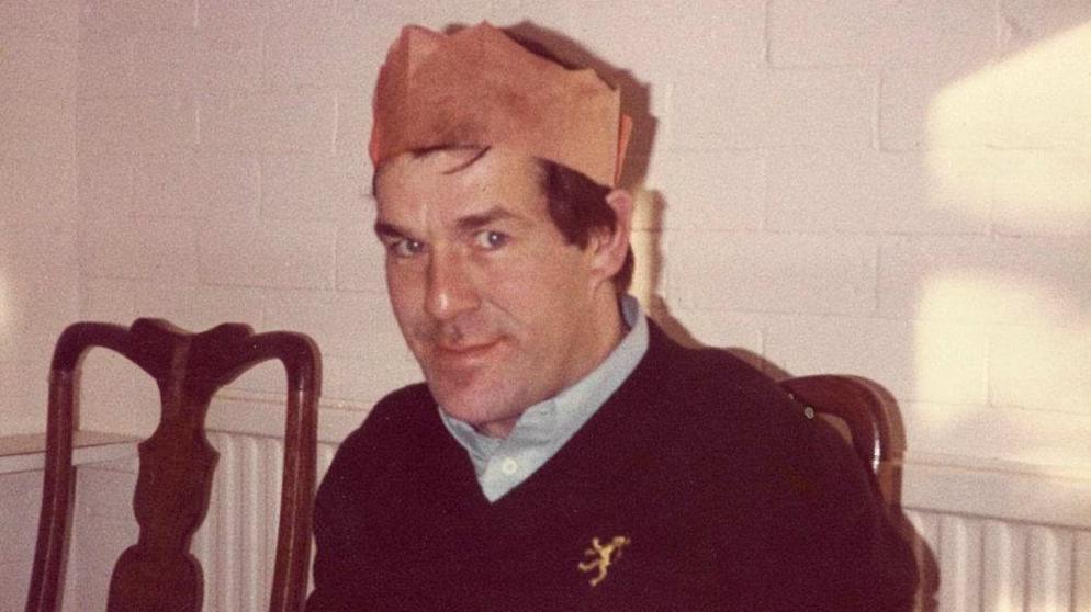 Photograph of Ronnie Lockwood, who wears a red paper party hat typically worn on Christmas day. He wears a blue shirt with a navy v neck jumper over the top of it. He has bright blue eyes and brown hair and smiles at the camera. 