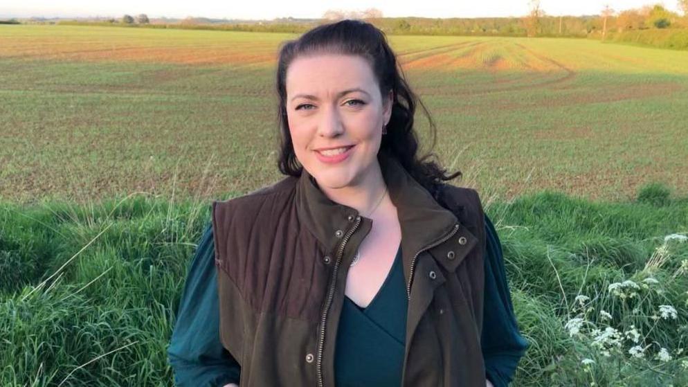 Alicia Kearns, MP for Rutland and Melton - A smiling woman in her 30s with long dark hair and blue eyes stood in a field.