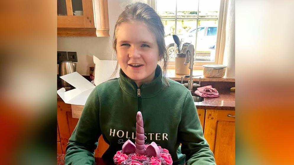 Anna Lowri Roberts celebrating her 11th birthday in January 2025. She is looking into the camera and smiling while holding a unicorn birthday cake in her kitchen