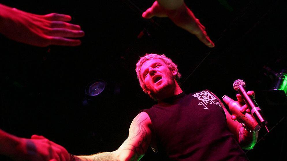 Mathew Pritchard, pictured from down below on a stage, which is partly lit. He holds a microphone and a bottle of beer in his right hand, and shakes the hand of someone with his left. Two hands can be seen reaching above his head. Pritchard wears a black vest and has blonde hair. His mouth is open as he appears to be speaking.