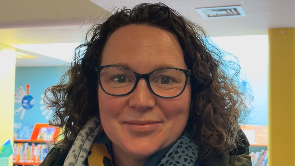 Laura John smiles at the camera. She wears black glasses and has short brown curly hair. She wears a scarf around her neck and a coat. 