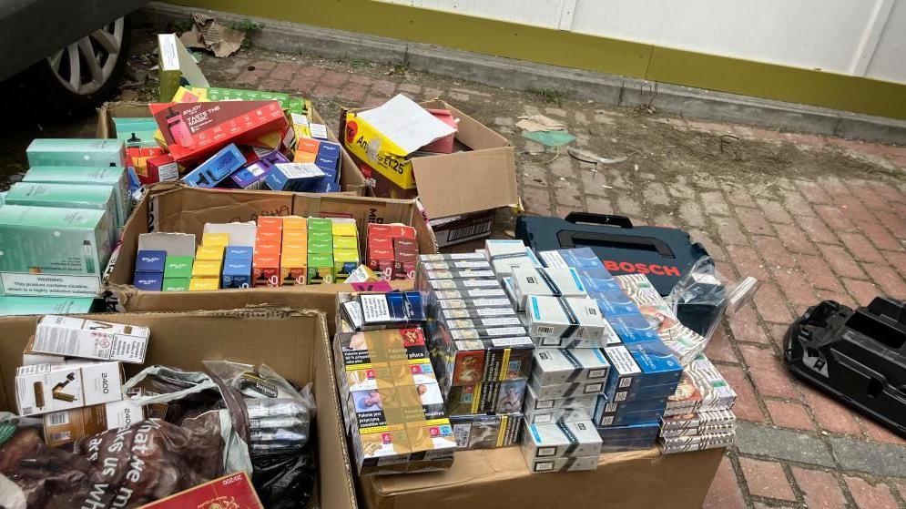 There are five boxes full of illegal vapes and tobacco on the ground on a drive way. They are stacked up against each other  and are open on the top, with vapes and tobacco spilling out of them. 