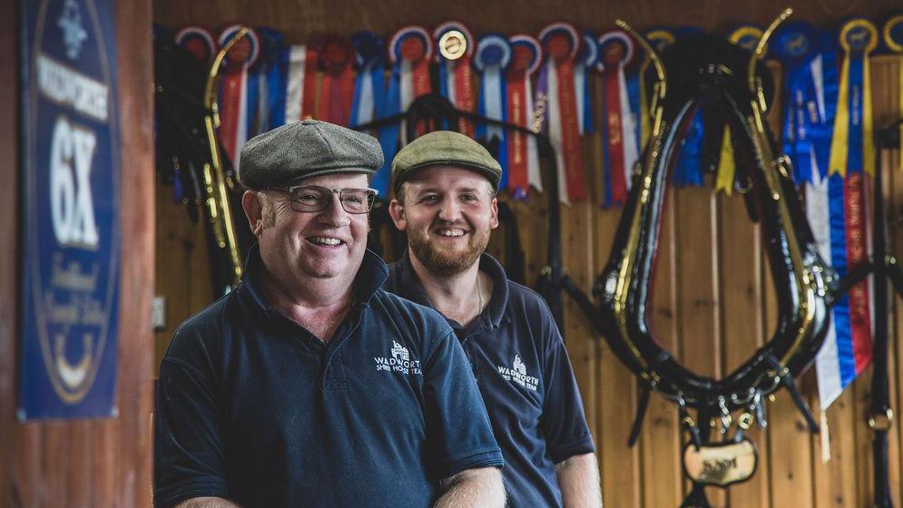 Martin and Callum Whittle smiling standing in front of a wooden wall covered in harnesses and rosettes. 