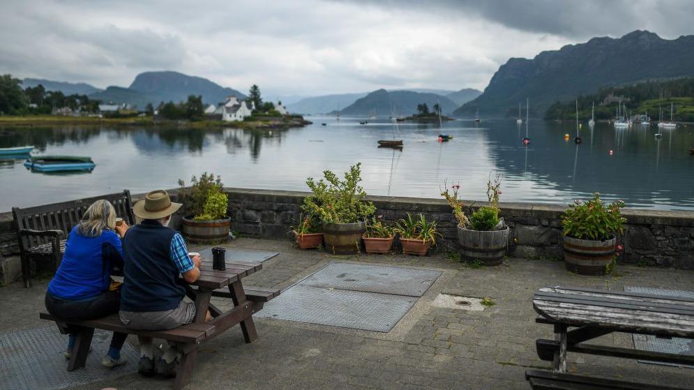 A couple sitting at a bench in Plockton. There are boats on the water and hills in the background