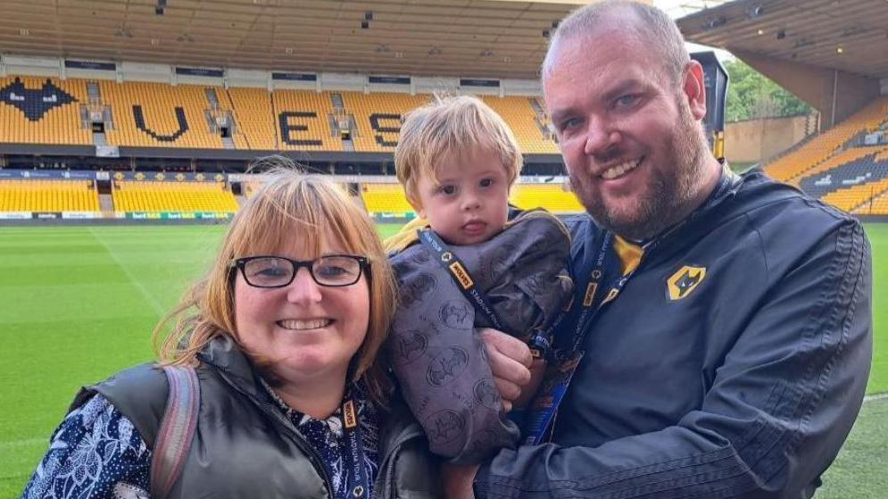 Katie and Aled holding Tomos and smiling at the camera, stood on the pitch at the Molineux football stadium in Wolverhampton. Katie is on the left and has a short light-brown hair in a bob, and is wearing a black gilet over a blue and white flowered top. Aled is on the right in a black top which has the Wolverhampton Wanderers logo on it. He has a dark brown stubbly beard. He holds Tomos who is sticking his tongue out at the camera.