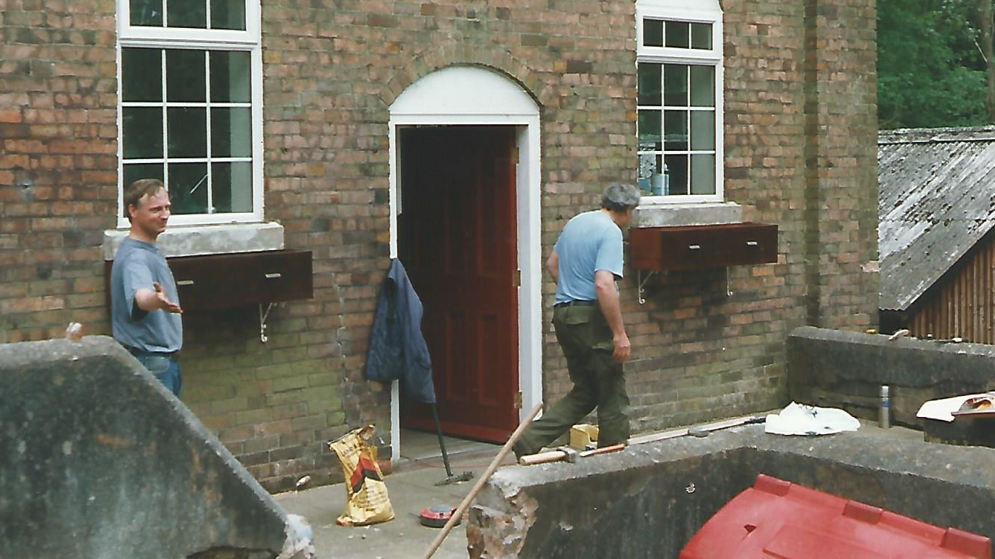 Two men are outside the chapel as renovations are carried out. Brooms, concrete bags and building equipment can be seen outside the chapel's entrance.
