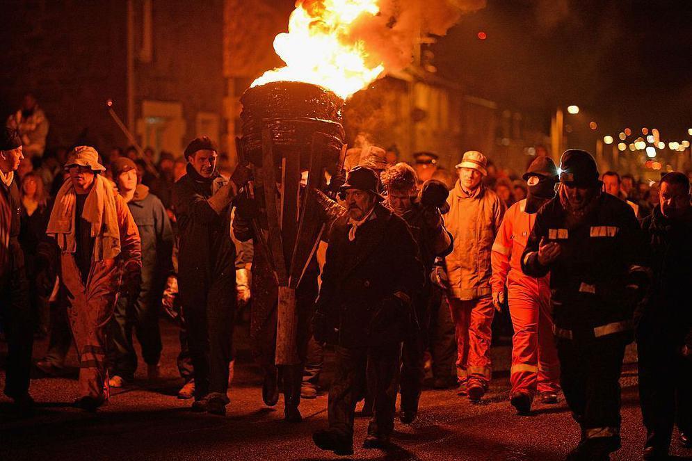The Clavie is a barrel of burning tar. Flames burn brightly from the top of the barrel and casts a group of men around it in an orange glow. The men, some of them wearing protective clothing, are take it turns to carry the Clavie down a street in Burghead.