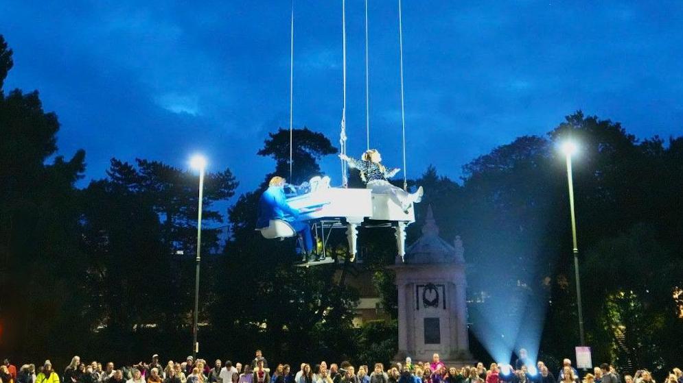 Performer sat on a white grand piano suspended in the air with a crowd watching.