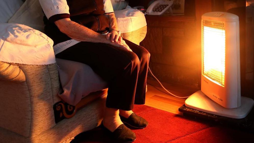 A pensioner wearing slippers, a brown and white woollen top and dark trousers sits in an armchair in front of an electric heater which is glowing orange.