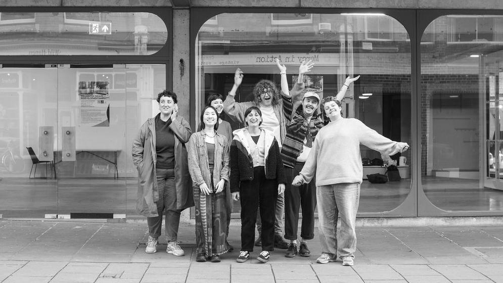 A black and white picture of a group of young people smiling and cheering outside a building. The building has ground to ceiling windows across its frontage.