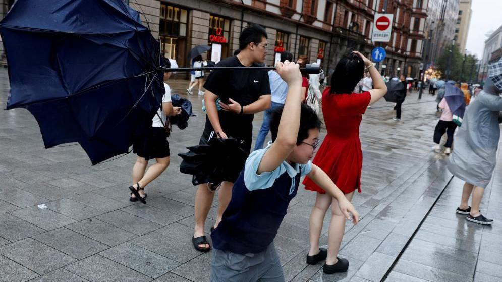 People holding umbrellas along a street in Shanghai. One boy is seen trying to hold on to an inverted umbrella as winds intensify