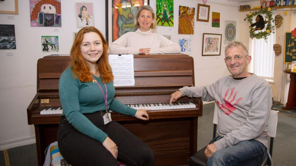 A man and two women beside a piano