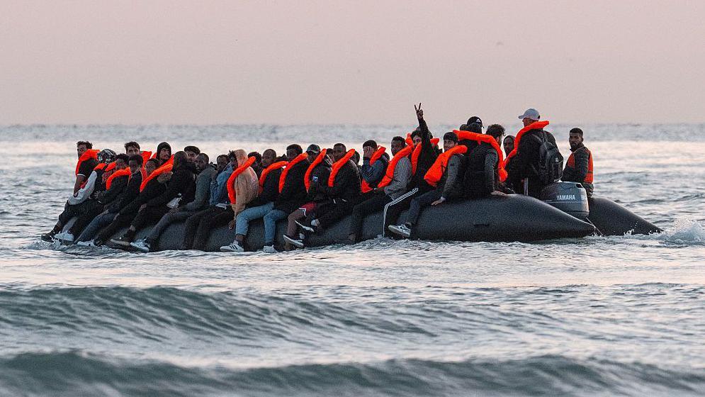 A rubber dinghy carrying migrants in the Channel. Men wearing bright orange lifejackets.