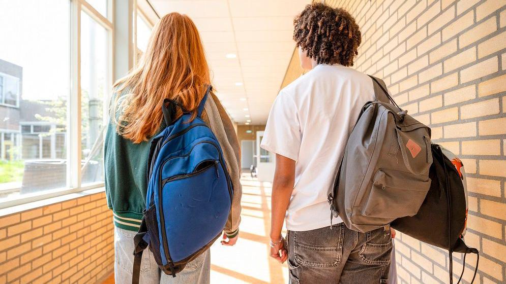 Two students walking ahead, facing away from the camera. One girl has red hair and is wearing a green jacket and a blue bakcpack, a boy is wearing a white t-shirt and is holding two grey backpacks, he has brown curly/afro hair. 