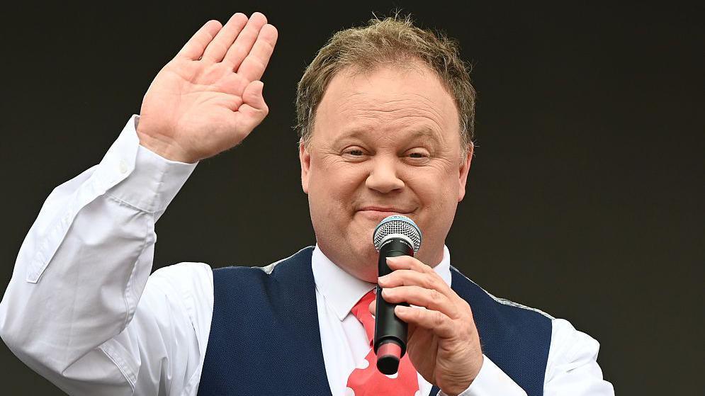 Justin Fletcher, waving with his right hand and a microphone in his left hand, is wearing a blue waistcoat, white shirt and a red tie with white dots on it. 