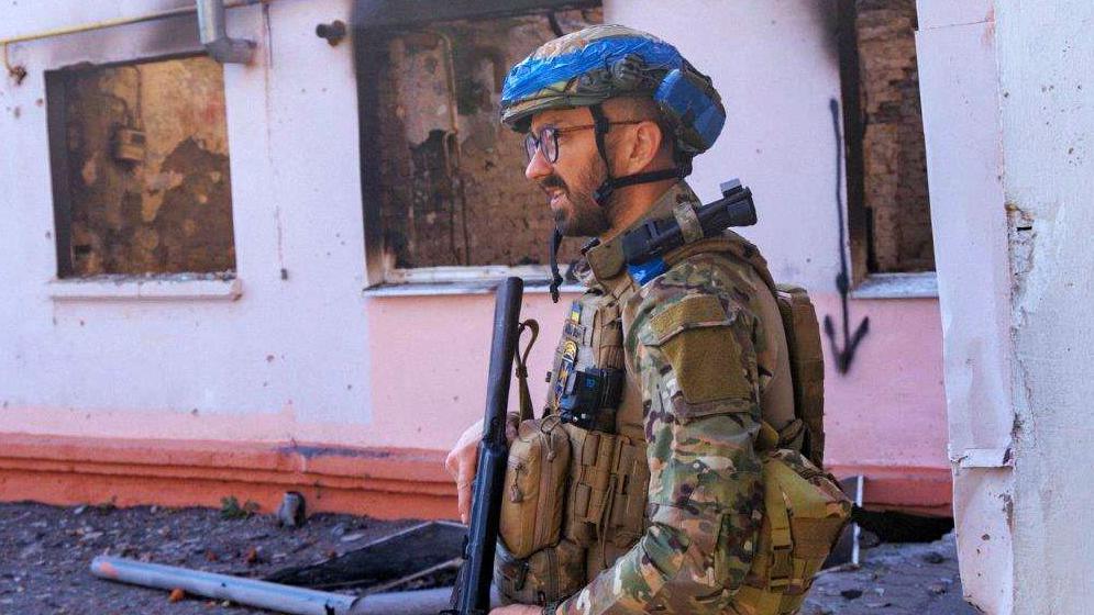 A Ukrainian military man holding a Kalashnikov rifle walks past a building that has blown out windows in September in Sudzha, Kursk Region, Russia.