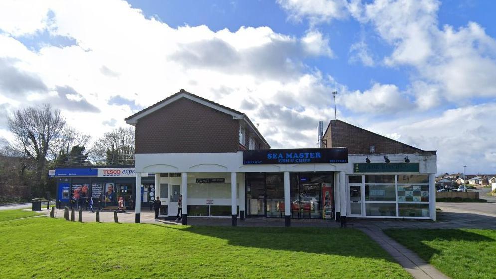 View of a row of retail units - red brick, mostly one storey, one has a level above. Grass in front, surrounded by a residential area. Sunny day.