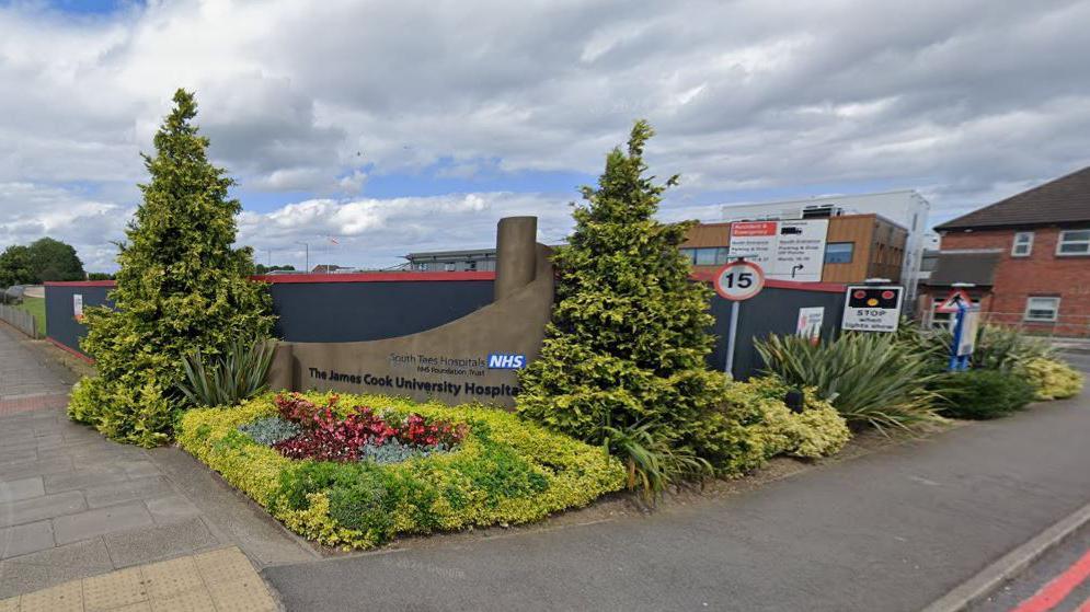 The entrance to the James Cook University Hospital. A wall displaying the hospital's name is surrounded by flowers and shrubs.