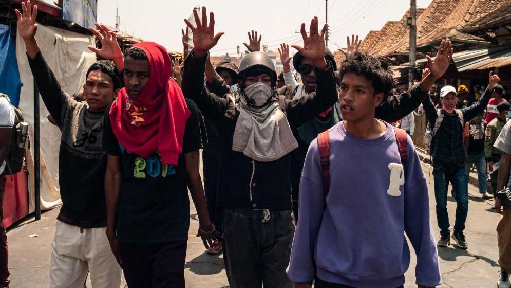 Protesters walk along with their hands in the air, some have their faces covered.